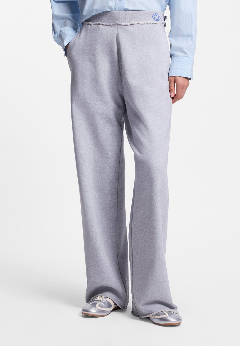 Άτομο που φοράει ανοιχτό γκρι παντελόνι φαρδιάς γραμμής, αθλητικά παπούτσια σε μεταλλικό ασημί και λευκό και ένα μπλε ριγέ πουκάμισο με κουμπιά, με τα χέρια στις τσέπες.