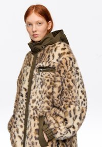 Mujer joven con cabello rojo que lleva un abrigo de piel con estampado de leopardo sobre una chaqueta verde oliva con capucha y bolsillo con cremallera en el pecho.