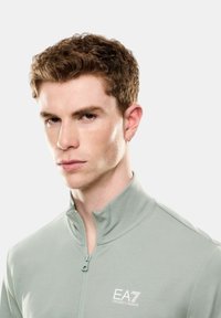 Giacca zip in verde chiaro con colletto alto, caratterizzata da un logo EA7 bianco sul petto e una texture liscia e aderente.