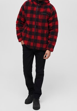 Hombre con chaqueta de forro polar a cuadros rojos y negros, pantalones negros y botas negras, de pie con una mano en el bolsillo.