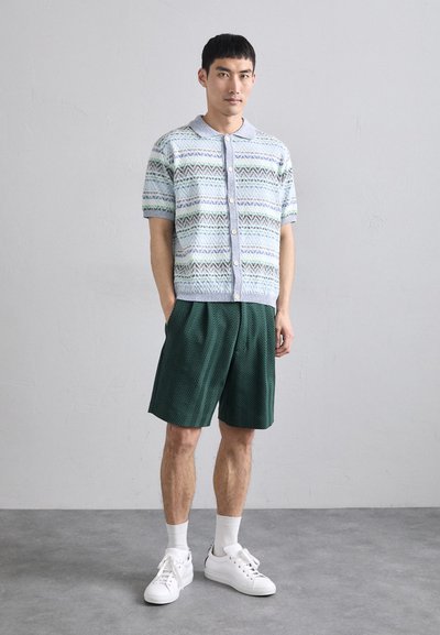 Knit short-sleeve shirt featuring a blue and green geometric pattern, paired with dark green herringbone-patterned shorts and white sneakers.
