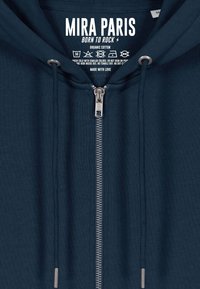 Haut à capuche zipé bleu marine avec fermeture éclair en métal argenté et cordons de serrage, présentant la marque "Mira Paris Born to Rock" et des instructions d'entretien à l'intérieur du col.