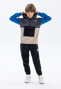Grijze, blauwe en beige hoodie met een voorzak; gecombineerd met zwarte joggers en witte sneakers. Glad materiaal, ontspannen pasvorm.