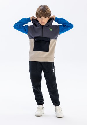 Kapuzensweatshirt in Grau, Blau und Beige mit einer Vordertasche; kombiniert mit schwarzen Jogginghosen und weißen Sneakers. Glattes Material, lockere Passform.