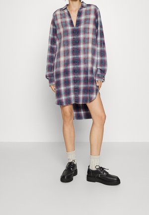 Kobieta ubrana w czerwoną i niebieską koszulę w kratę o długim rękawie, w białych skarpetkach i czarnych butach sznurowanych, stojąca na jednolitym tle.