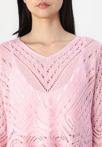 Rosa stickad tröja med V-ringning, med ett geometriskt spetsmönster och öppna stygn. Mjuk textur och avslappnad passform.