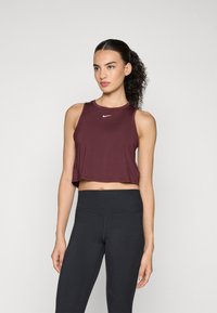 Woman with curly ponytail wearing a sleeveless maroon crop top and high-waisted black leggings, standing against a plain background.