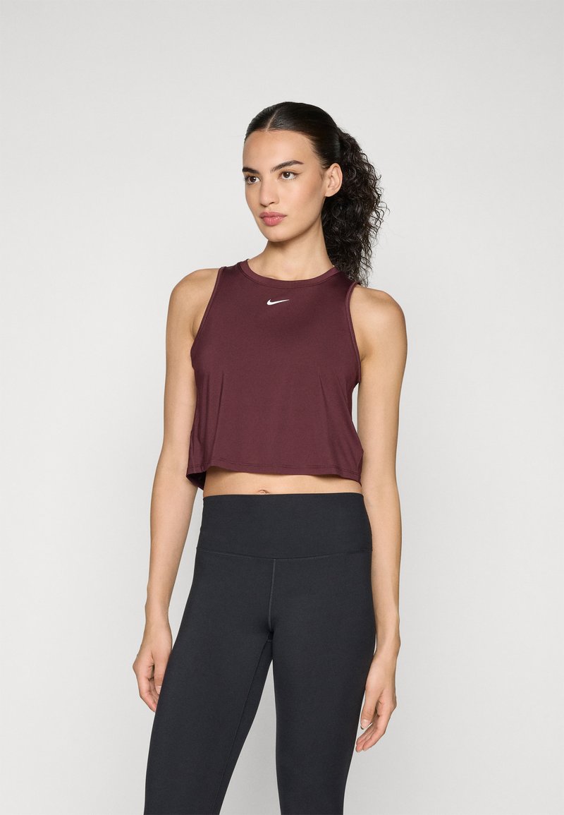 Woman with curly ponytail wearing a sleeveless maroon crop top and high-waisted black leggings, standing against a plain background.