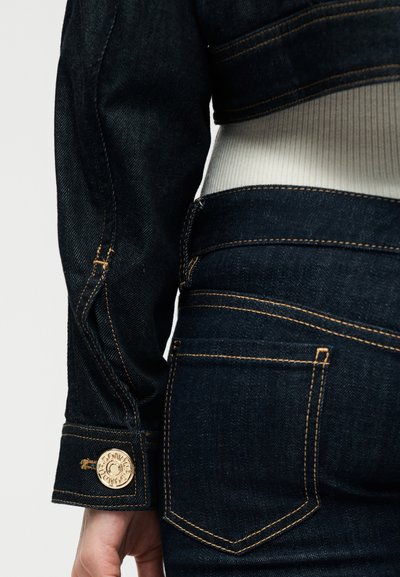 Κλείσιμο εικόνας ατόμου που φορά σκούρο μπλε τζιν μπουφάν και τζιν με χρυσή ραφή και κουμπί, με εμφανές λευκό ριγωτό πουκάμισο από κάτω.