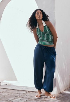 Göndör hajú nő zöld csíkos pántnélküli felsőben, laza tengerészkék nadrágban és fehér szandálban áll egy fehér fal előtt, amelyen ív található.