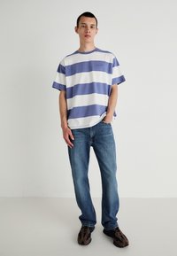 T-shirt de manga curta riscada em branco e roxo claro, jeans de denim e ténis castanhos. Corte descontraído com decote redondo e mangas largas.