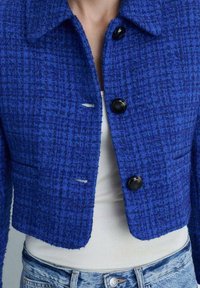 Giacca cropped blu realizzata in tessuto strutturato, con colletto, bottoni frontali e tasche laterali. Bottoni neri accentuano il design.