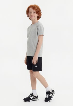 Rødharret gutt står sidelengs og har på seg en grå t-skjorte, svarte New Balance-shorts, hvite sokker og svarte New Balance-sneakers.