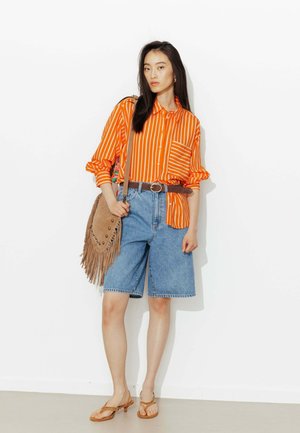 Femme portant une chemise rayée orange et blanche, un short en jean longueur genoux, une ceinture marron, un sac à franges beige à l'épaule et des sandales beige à talons, debout.