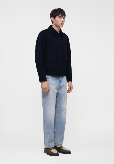 Homme portant une veste noire, un jean bleu clair délavé et des chaussures en cuir tressé marron et beige, debout devant un fond clair uni.