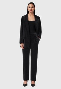 Donna che indossa un blazer di velluto nero e pantaloni abbinati, un top senza spalline a costine nero e décolleté a punta neri, in piedi davanti a uno sfondo grigio.