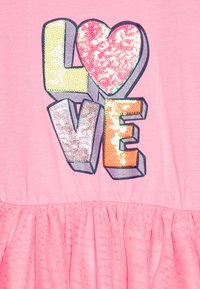 Ροζ μπλουζάκι με τη λέξη "ΑΓΑΠΗ" σε πολύχρωμες παγιέτες. Διαθέτει μια γυ glitter καρδιά, επικαλυμμένη με μια υφασμάτινη φούστα από τούλι στο κάτω μέρος.