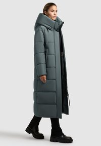 Cappotto lungo imbottito in verde teal tenue con colletto alto e zip frontale. Presenta un'imbottitura orizzontale, tasche laterali e una vestibilità comoda.