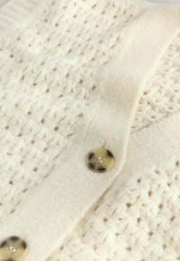Cardigan en tricot couleur crème avec un motif texturé, des poignets côtelés et deux grands boutons transparents.