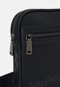 Černá crossbody taška s texturovanou tkaninou a hladkými koženými akcenty, dvojitými zipy s logem na vytahovačích a odolným popruhem.