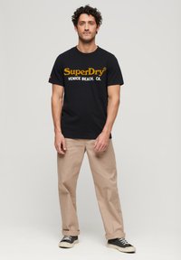 Czarna bawełniana koszulka z logo "SuperDry" w kolorze pomarańczowym, w połączeniu z beżowymi spodniami i czarnymi sneakersami z białymi podeszwami.