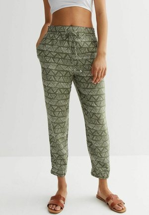 Žena v olivově zelených teplákových kalhotách s bílým geometrickým potiskem, bílém krátkém topu a hnědých nazouvacích sandálech, stojící s rukama v kapsách.