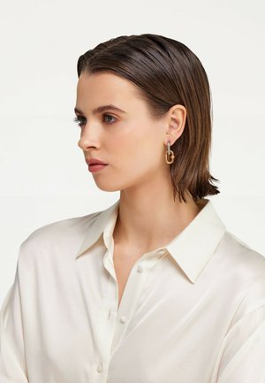 Boucles d'oreilles créoles en or et noir avec un design connecté. Texture lisse avec une finition brillante, portées par une personne portant une blouse de couleur claire.