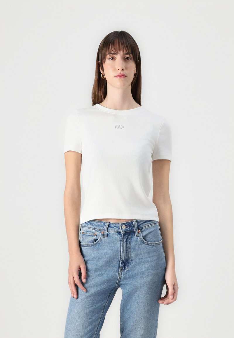 Tricou alb cu mânecă scurtă, cu o textură ribbed și logo-ul brodat "GAP" pe piept, asortat cu blugi din denim de un albastru deschis.