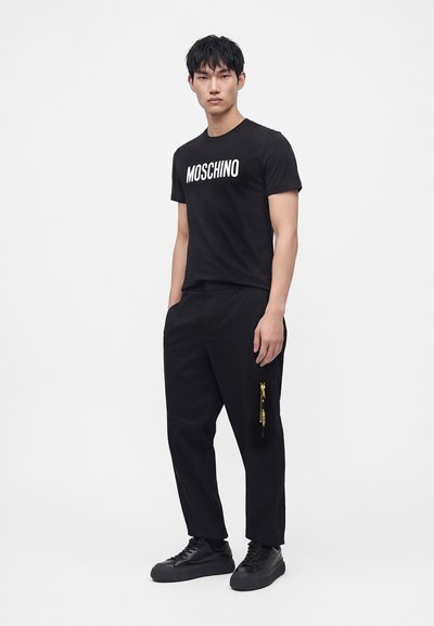 Νέος άνδρας που φοράει μαύρο T-shirt Moschino, μαύρο παντελόνι με φερμουάρ στο πλάι και μαύρα αθλητικά παπούτσια, σε λευκό φόντο.