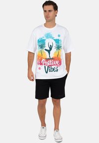 Wit katoenen T-shirt met een kleurrijke afbeelding van een danser, palmbomen en de tekst "Positive Vibes". Gecombineerd met zwarte shorts en sneakers.