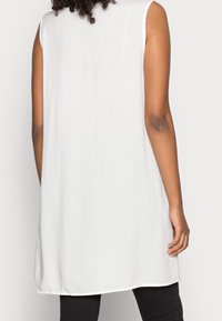 Tunique blanche sans manches à coupe ample, fabriquée en tissu doux, avec un ourlet arrondi et des fentes latérales pour faciliter les mouvements.