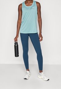 Lichtblauw mouwloos sporttanktop met een ronde halslijn, gecombineerd met donkerblauwe leggings. De persoon houdt een zwarte waterfles vast.