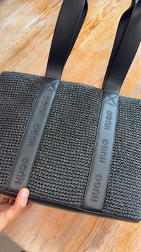 Sac fourre-tout tissé noir avec deux bretelles en cuir embossées du mot "HUGO" tenu au-dessus d'une surface en bois.