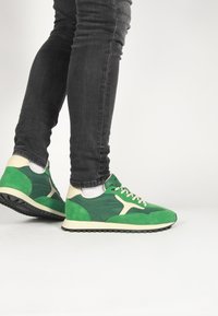 Zelené semišové a kožené tenisky s krémovými akcentmi, ktoré majú štruktúrovanú podrážku, šnurovanie na prednej strane a kontrastné prekrytia.