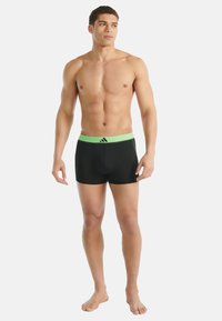 Černé boxerky s elastickým zeleným pasem s logem Adidas. Materiál vypadá hladce, pružně a přiléhavě.