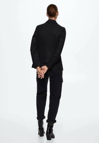 Zwarte op maat gemaakte blazer met een textuurafwerking, gedragen over donkere cargobroeken en enkellaarsjes. Het onderwerp heeft de handen achter de rug clasped.