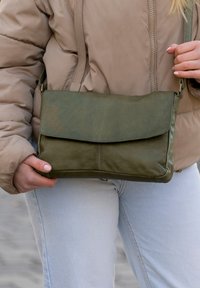 Olivnozeleni usnjen nahrbtnik preko telesa z zložljivim pokrovom, gladko teksturo in trdnimi šivi, držan z eno roko ob svetli obleki.