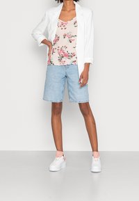 Donna con un blazer bianco, un top floreale rosa, pantaloni di jeans leggermente blu e sneakers bianche con dettagli rosa, in piedi con una mano appoggiata all'anca.