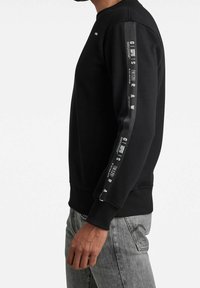 Sudadera negra con puños y dobladillo acanalados. Presenta una franja brillante en la manga con texto y logos blancos. Diseño de cuello redondo.