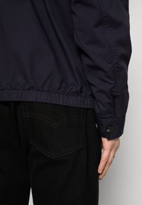 Marinejakke med elastisk kant og mansjetter, som kombinerer en jevn tekstur med funksjonelle knapper. Brukt over svarte bukser med et baklomme-detalj.