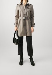 Trench coat beige con vita cinturata, ampio colletto e bottoni; abbinato a pantaloni neri a vita alta e stivali neri alla caviglia; in mano una borsa nera.