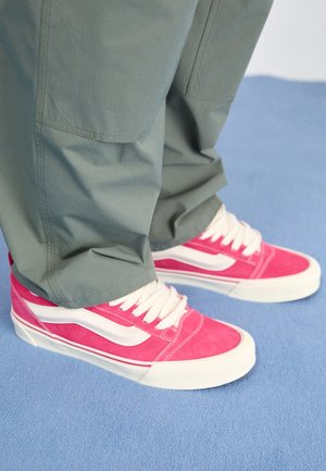Baskets lacées rose vif et blanc portées avec un pantalon vert ample, debout sur un sol recouvert d'un tapis bleu clair.
