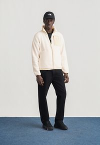 Άνδρας που στέκεται σε μπλε χαλί φορώντας μπεζ φλις μπουφάν, μαύρο παντελόνι, μαύρα αθλητικά παπούτσια και μαύρο καπέλο με εμφανές λογότυπο.