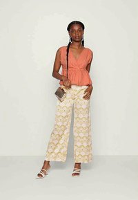 Top texturizado em pêssego com decote em V e cintura peplum combinado com calças largas cremes apresentando um padrão de concha dourada. Os acessórios incluem uma bolsa.