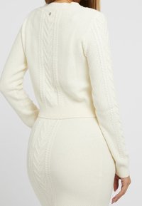 Biały sweter z dzianiny o teksturowanym ciele i detalach w splot warkoczowy na rękawach. Posiada prążkowany dół oraz szew centralny z tyłu.
