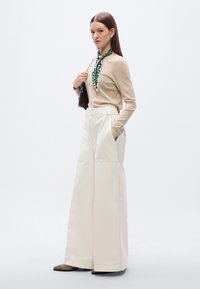 Ευρύχωροι κρεμ παντελόνια, μπεζ πλεκτό τοπ, πράσινο φουλάρι με σχέδια δεμένο γύρω από τον λαιμό και καφέ παπούτσια με μυτερή μύτη. Ασημένια μεταλλικά στοιχεία σε μαύρη τσάντα χειρός.