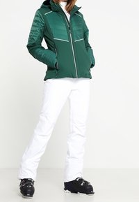 Veste isolante verte avec fermeture éclair à l'avant, accents blancs contrastants et manches texturées, accompagnée d'un pantalon de neige blanc et de chaussures de ski noires.