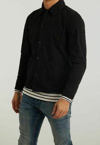 Zwarte button-up jas met een borstzak, lange mouwen en een licht gestructureerde stof, gelaagd over een gestreept wit en zwart shirt.