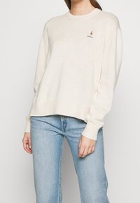 Cremfarbener Strickpullover mit gerippten Manschetten und Saum, versehen mit einem kleinen gestickten Logo auf der Brust. Kombiniert mit hellblauen Jeans.