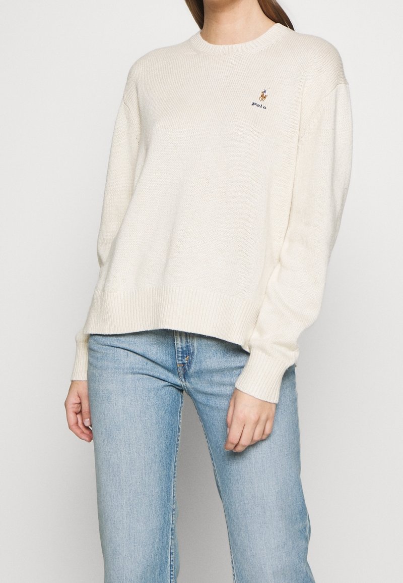Cremfarbener Strickpullover mit gerippten Manschetten und Saum, versehen mit einem kleinen gestickten Logo auf der Brust. Kombiniert mit hellblauen Jeans.